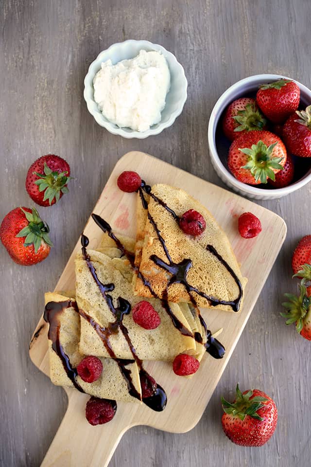 Chopping board with folded Parsi crepes surrounded by bowls with whipped cream and strawberries
