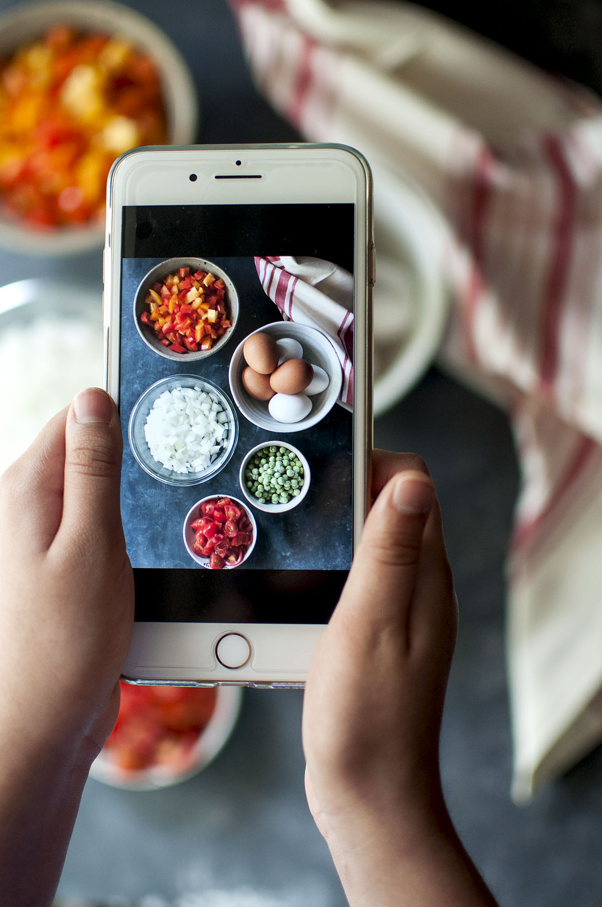 Hand holding a phone with a photo of onions, bell peppers, tomatoes, green peas and eggs 