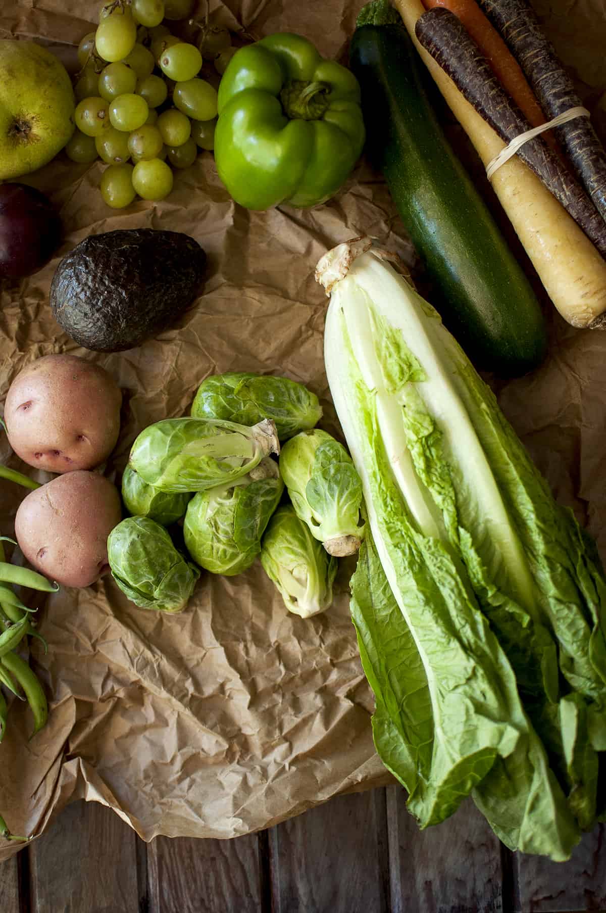 Brown paper with lettuce, brussel sprouts, potatoes, avocado, pepper, zucchini, carrot, grapes and pear