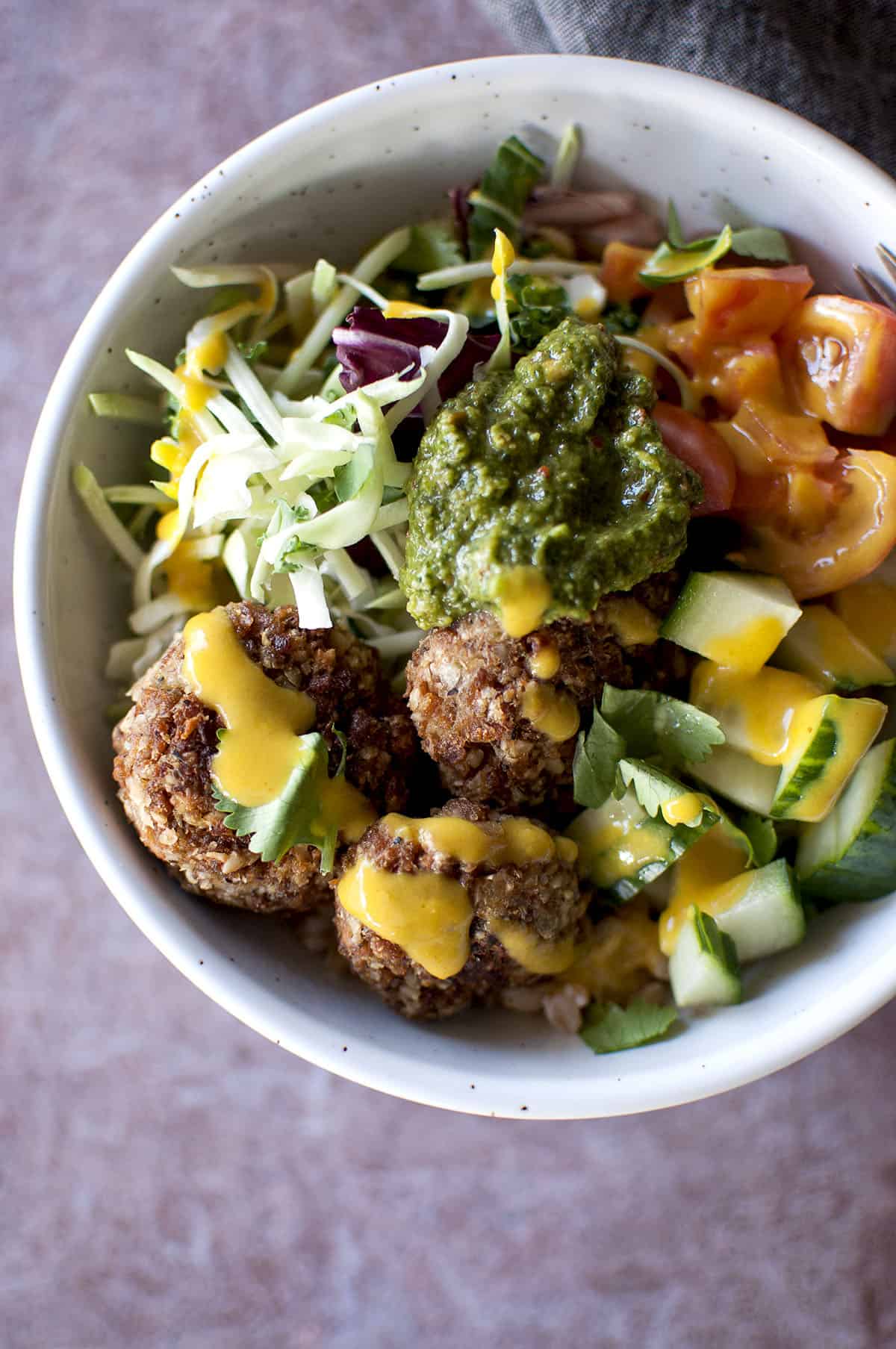 Grey bowl with meatless meatballs, chopped cucumbers, tomatoes, salad greens, green chutney and amba sauce