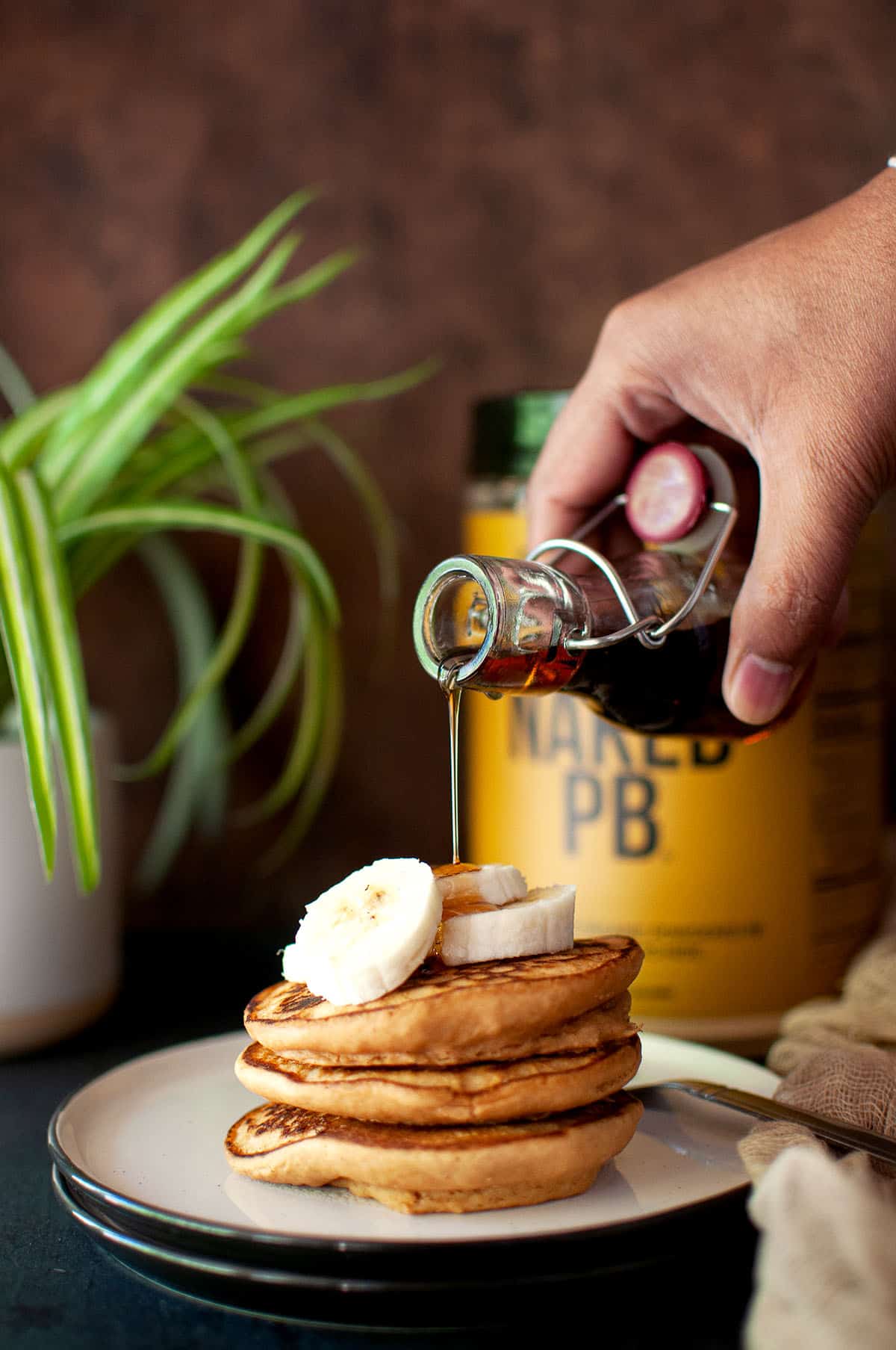 Hand pouring maple syrup over a stack of peanut butter pancakes.